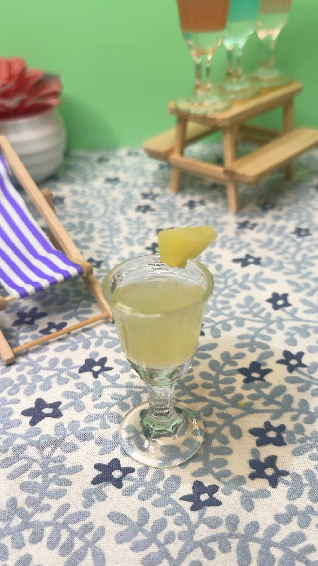 500114499_18499460314027811_8206755621951528713_n Mini pineapple cocktail on a floral tablecloth, with a striped deck chair and tiny picnic table holding colorful drinks.