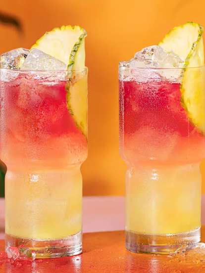 Two Malibu Bay Breeze cocktails served in highball glasses with a pineapple slice standing on a orange tray