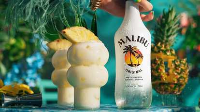 Malibu Piña Colada cocktails in curvy glasses with creamy white drink and pineapple wedge garnish, condensation on glass, Malibu Original bottle held beside, tropical turquoise background with bright summer lighting.
