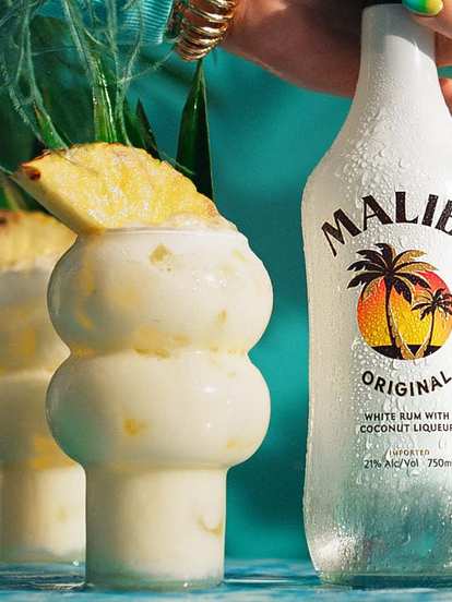 Malibu Piña Colada cocktails in curvy glasses with creamy white drink and pineapple wedge garnish, condensation on glass, Malibu Original bottle held beside, tropical turquoise background with bright summer lighting.