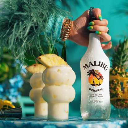 Malibu Piña Colada cocktails in curvy glasses with creamy white drink and pineapple wedge garnish, condensation on glass, Malibu Original bottle held beside, tropical turquoise background with bright summer lighting.