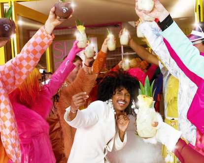 A diverse group of people in vibrant outfits joyfully raising their drinks in celebration.