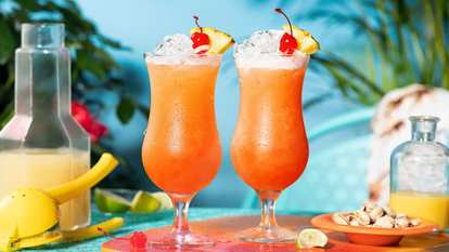 Malibu Bahama Mama — two tall orange tropical cocktails topped with crushed ice, pineapple slices, and cherries. The bright setup with citrus tools and nuts evokes a sunny, beach‑bar vibe.