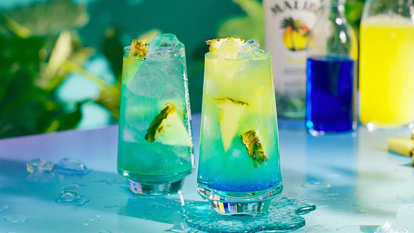 Two Malibu Blue Hawaiian cocktails garnished with multiple pineapple slices standing on a blue table in front of a Malibu Original bottle