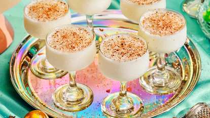 A festive tray of Malibu Coquito served in coupe glasses, each filled with a creamy coconut rum cocktail topped with a dusting of nutmeg. The iridescent serving tray reflects warm holiday tones, creating a cozy, celebratory atmosphere perfect for seasonal gatherings.