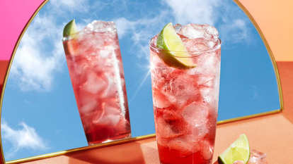 Two tall glasses filled with pink Malibu Cranberry Lemonade over ice, garnished with fresh lime wedges, one reflected in a mirror against a bright sky-blue and coral backdrop.