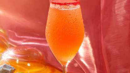 Chilled Malibu Fizz cocktail in a flute glass with a light pink to orange gradient, fine bubbles and condensation, rim dipped in red sugar, set against a vibrant, glossy pink background.
