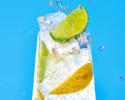 Malibu Soda cocktail in a tall glass with clear sparkling drink, ice cubes, and lime wedges, water droplets mid‑air showing splash motion, set against a bright blue background for a fresh summer feel.