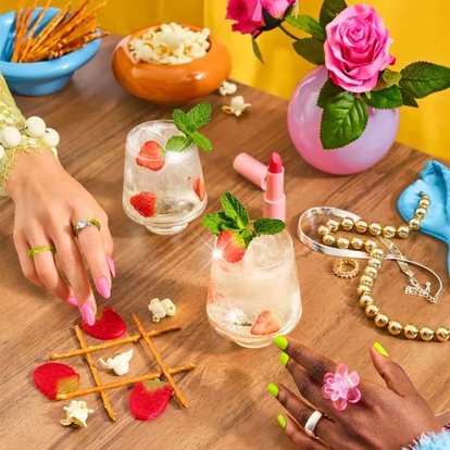 Malibu Strawberry Spritz cocktails on wooden table with ice, mint, and strawberry slices, surrounded by playful props, jewelry, popcorn, and bright pink roses in a lively setting.