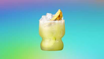 A short, curved glass of Malibu Kiwi Dream filled with ice cubes and slices of fresh kiwi, topped with a pineapple wedge garnish. The light green coconut rum cocktail glistens with condensation against a vibrant gradient background, evoking a refreshing, tropical summer vibe with a fruity twist.