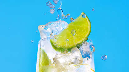 Malibu Lemon‑Lime Soda cocktail close‑up with lime slices and ice cubes, water droplets splashing around the glass against a bright blue background, capturing freshness and motion.