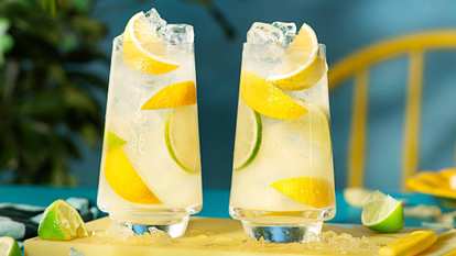 Malibu Lemonade cocktails in tall glasses with clear drink, ice cubes, lemon and lime slices, placed on a yellow surface with scattered ice against a turquoise tropical background.