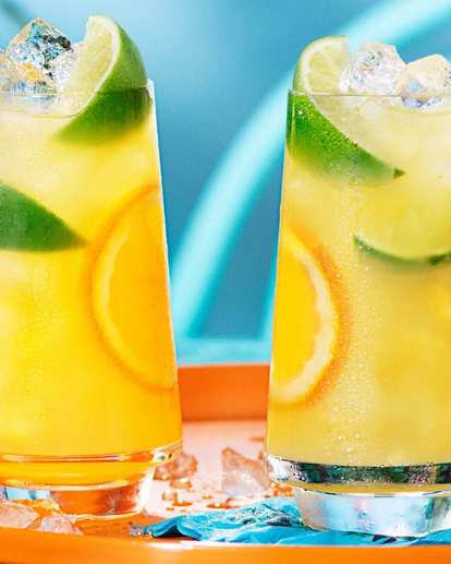 Two tall glasses of Malibu and orange juice filled with ice, lime wedges, and orange slices on a bright summer tray. A tropical coconut rum cocktail with citrus notes in a sunny outdoor setting.
