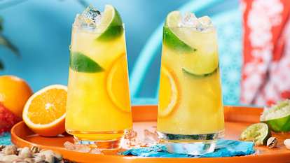 Two tall glasses of Malibu and orange juice filled with ice, lime wedges, and orange slices on a bright summer tray. A tropical coconut rum cocktail with citrus notes in a sunny outdoor setting.