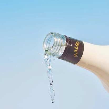 Malibu Original bottle pouring clear liquid mid‑air against bright blue sky, sparkling reflections highlighting freshness and clarity in a minimal tropical composition.