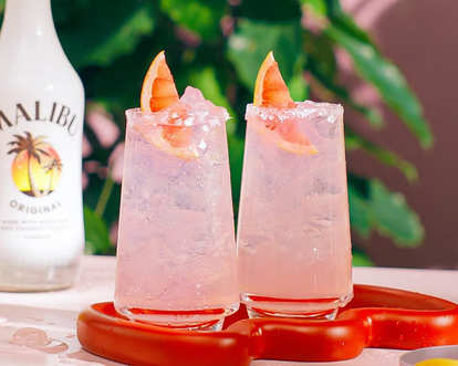 Two Malibu Paloma cocktails in tall glasses with light pink drink, ice cubes, grapefruit slices, and salted rims, Malibu Original bottle blurred in background.