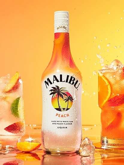 A Malibu Peach bottle centered between two peach cocktails. The left glass is garnished with mint and a peach slice, while the right tall glass features ice, peach wedges, and a strip of citrus peel. The soft pink sky background adds a warm, summery, tropical feel.