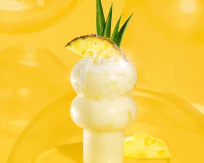 Malibu  Piña Colada cocktail in curvy glass with creamy yellow drink, ice, pineapple slice, and green leaves garnish, glowing against a vibrant yellow background.