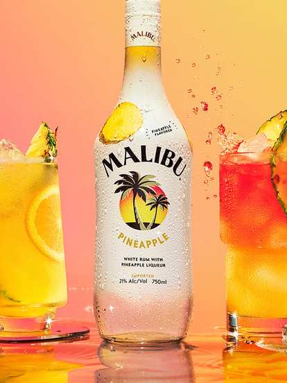 A Malibu Pineapple bottle placed between two tropical cocktails. On the left, a bright yellow drink with ice, lemon slice, and pineapple wedge; on the right, a layered red‑and‑yellow cocktail with ice and pineapple garnish. The orange‑to‑pink background and droplets create a fresh, sunny, beach‑party vibe.