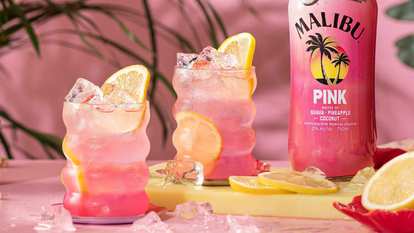 Malibu Pink Lemonade cocktails in highball glasses with pink layered drink, ice cubes, and lemon slices, placed beside a Malibu Pink bottle on a pastel surface with scattered ice and sliced lemons.
