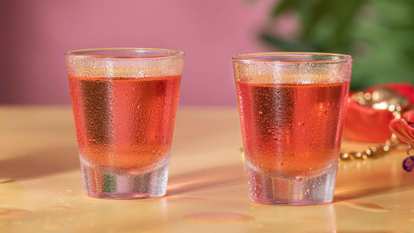 Two Malibu Pink Shot cocktails in clear shot glasses with chilled pink liquid and condensation, standing on a light surface with pink backdrop and golden decorative beads in the background.