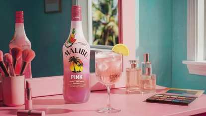 Malibu Pink Spritz cocktail in a stemmed glass with ice and a lemon slice garnish, placed beside a Malibu Pink bottle on a pink vanity with makeup brushes, perfume bottles, and an eyeshadow palette in the background.