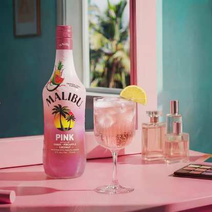 Malibu Pink Spritz cocktail in a stemmed glass with ice and a lemon slice garnish next to a Malibu Pink bottle on a pink surface, with perfume bottles and a window showing palm trees in the background.