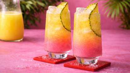 Two Malibu Pink and Pineapple Juice cocktails in tall glasses with pineapple slices and crushed ice on red coasters against a pink tropical background.