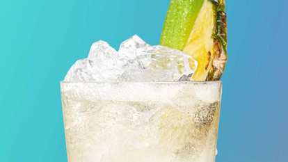 Malibu Sparkling Piña Colada cocktail close‑up with light creamy drink, ice cubes, and pineapple wedge garnish, condensation on the glass against a turquoise blue background.