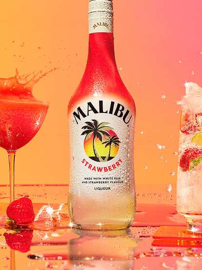 A Malibu Strawberry bottle placed between two vibrant strawberry cocktails. On the left, a frozen strawberry drink splashes upward in a stemmed glass; on the right, a tall glass with ice, sliced strawberries, and lime wedges sparkles in the light. The warm orange‑to‑pink background enhances the tropical, fruity energy.