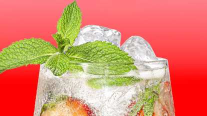 Malibu Strawberry Spritz cocktail close‑up with clear sparkling drink, ice cubes, strawberry slices, and mint leaves, condensation on the glass against a bright red background.