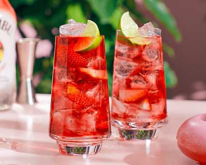 Malibu Strawberry Woo Woo cocktails in tall glasses with bright red drink, ice cubes, sliced strawberries, and lime wedge garnish, placed on a light pink surface with blurred green foliage in the background.