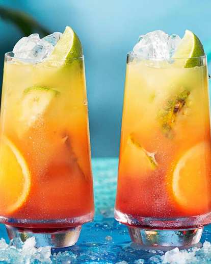 Two tall glasses of Malibu Sunrise with ice, lime wedges, and orange slices. The drink shows a vivid gradient from deep red to bright yellow, set on a turquoise table with scattered ice and citrus for a fresh, tropical look.