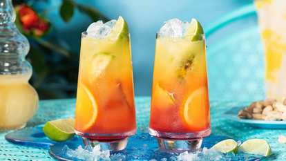 Two tall glasses of Malibu Sunrise with ice, lime wedges, and orange slices. The drink shows a vivid gradient from deep red to bright yellow, set on a turquoise table with scattered ice and citrus for a fresh, tropical look.