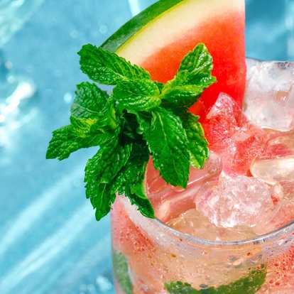 Malibu Watermelon Mojito cocktail in a clear glass with pink sparkling drink, ice cubes, fresh mint leaves, and a watermelon wedge garnish, condensation on the glass, isolated on a bright blue background.