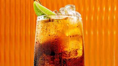 Malibu & Cola cocktail in a tall glass with dark amber drink, ice cubes, and a lime wedge garnish, condensation on the glass against a warm orange background.