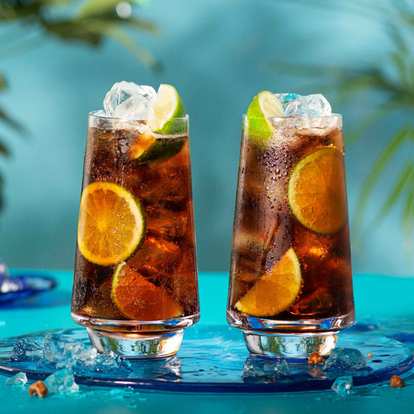 Malibu & Cola cocktail in a tall glass with dark amber drink, ice cubes, and a lime wedge garnish, condensation on the glass against a warm orange background.