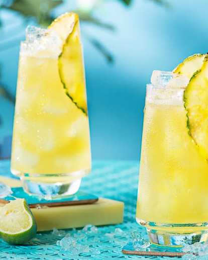 Two tall glasses of Malibu & Pineapple Juice with ice and pineapple wedges on a turquoise table. Fresh pineapple and lime pieces surround the drinks, with a bottle of juice and snacks in the background, creating a bright, summery atmosphere.