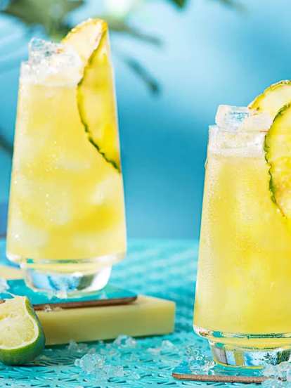 Two tall glasses of Malibu & Pineapple Juice with ice and pineapple wedges on a turquoise table. Fresh pineapple and lime pieces surround the drinks, with a bottle of juice and snacks in the background, creating a bright, summery atmosphere.