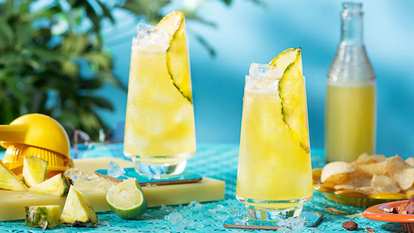 Two tall glasses of Malibu & Pineapple Juice with ice and pineapple wedges on a turquoise table. Fresh pineapple and lime pieces surround the drinks, with a bottle of juice and snacks in the background, creating a bright, summery atmosphere.