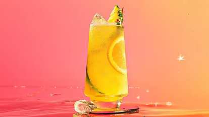 A single glass of Malibu & Pineapple Juice with ice, an orange slice, cucumber ribbon, and pineapple wedge garnish. Set against a pink‑to‑orange gradient background that glows with tropical warmth and sparkle.