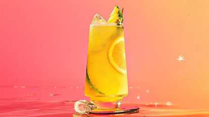 A single glass of Malibu & Pineapple Juice with ice, an orange slice, cucumber ribbon, and pineapple wedge garnish. Set against a pink‑to‑orange gradient background that glows with tropical warmth and sparkle.