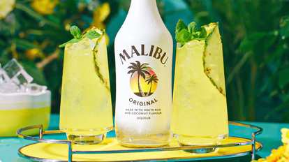A chilled Malibu Original bottle centered between two glasses of Malibu & Pineapple Juice. Each glass is garnished with pineapple and mint, placed on a yellow tray with green foliage in the background for a fresh, tropical vibe.