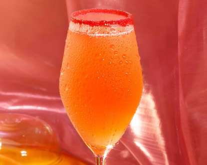 Malibu Baubelini cocktail in a stemmed glass with bright orange drink, red sugar rim, and condensation droplets, glowing softly against a pink satin background.