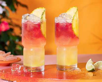 Two Malibu Bay Breeze cocktails in tall glasses with layered yellow‑to‑red gradient, ice cubes, and pineapple wedges, placed on orange surface with scattered ice and lime slices.