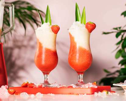Malibu Miami Vice cocktails in tall hurricane glasses with layered red and white frozen drink, strawberry and pineapple leaf garnish, tropical background with blender and plants, scattered ice on surface.