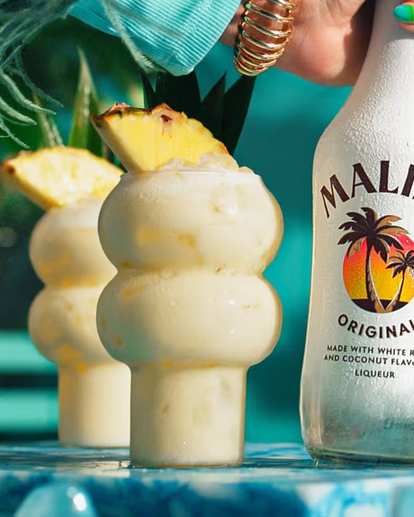 Malibu Piña Colada cocktails in curvy glasses with creamy white drink and pineapple wedge garnish, condensation on glass, Malibu Original bottle held beside, tropical turquoise background with bright summer lighting.