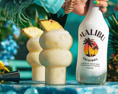 Malibu Piña Colada cocktails in curvy glasses with creamy white drink and pineapple wedge garnish, condensation on glass, Malibu Original bottle held beside, tropical turquoise background with bright summer lighting.