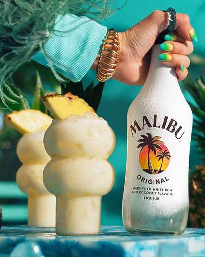 A hand holding a bottle of Malibu and two glasses of Pina colada on the side