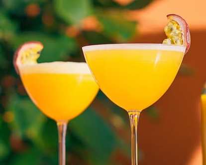 Porn Star Martini cocktails in coupe glasses with smooth yellow drink and frothy top, garnished with halved passion fruit, set against blurred green foliage and warm orange background for a tropical glow.